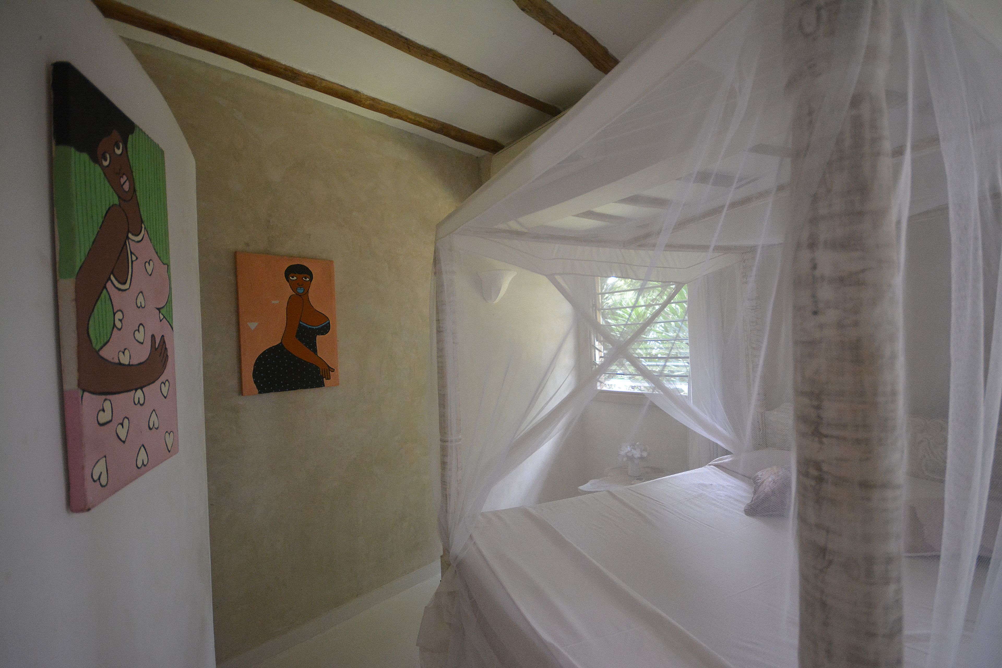 A room with a bed under a mosquito net, a painting on the wall, and a window with natural light.
