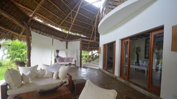 A covered outdoor seating area with a thatched roof, white cushioned furniture, and a stone floor.