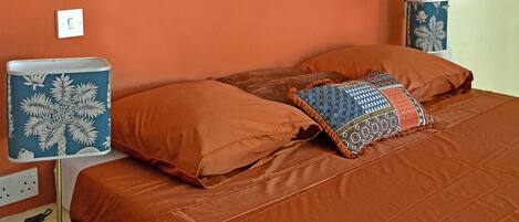 A bedroom with an orange wall, a bed with orange bedding, a bedside table with a lamp, and a power outlet.