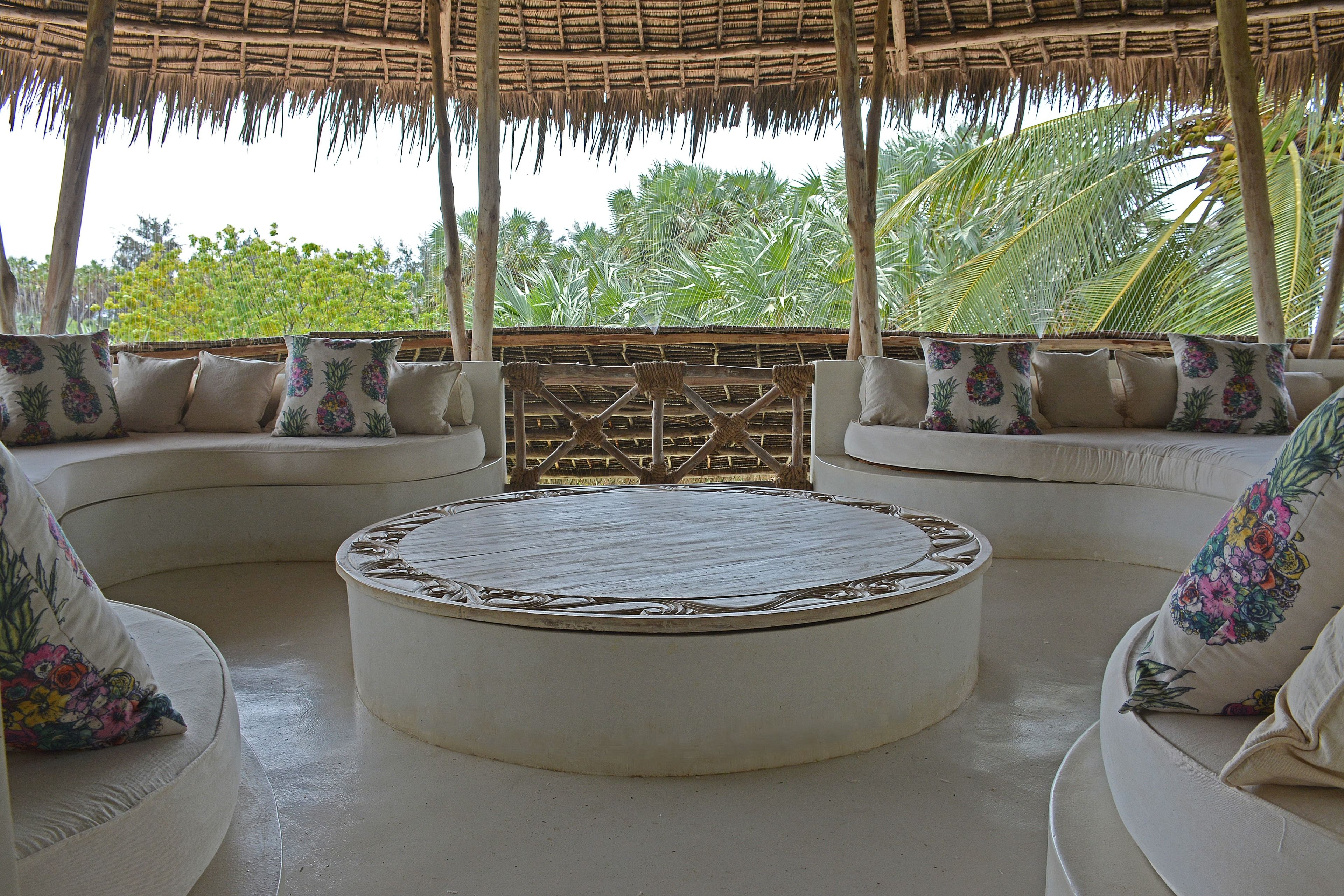 A round seating area with a woven ceiling, surrounded by cushioned sofas with floral pillows, under a thatched roof.