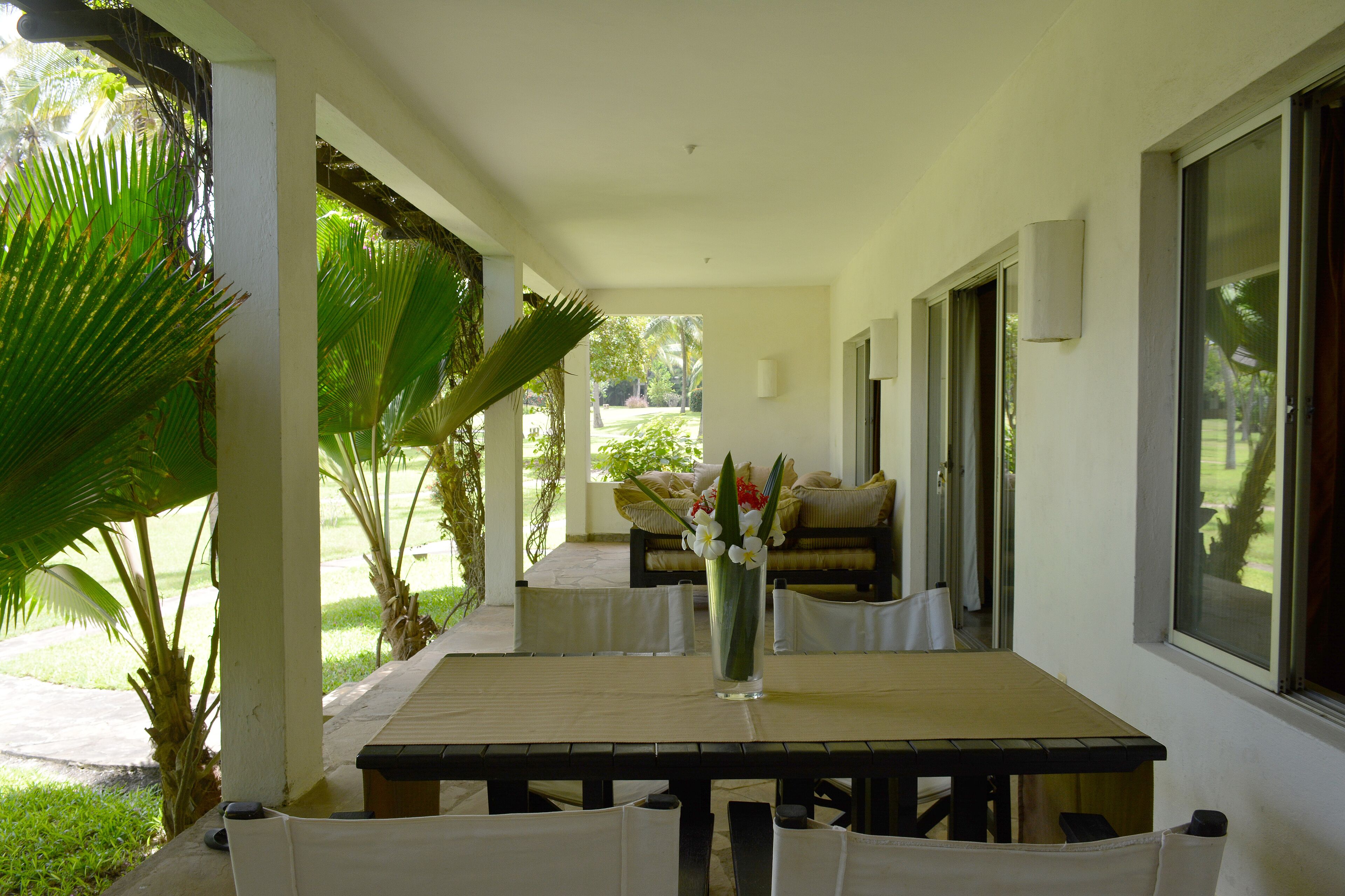 A covered patio with a wooden table and chairs, a vase of flowers, and a view of the garden.