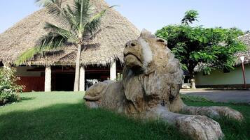 Una estatua de león de piedra en un área cubierta de césped, con un edificio de techo de paja y palmeras al fondo.