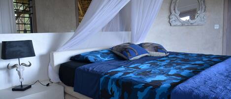Un dormitorio con cama con dosel, una mesita de noche con lámpara, un espejo en la pared y una ventana con barrotes.