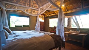 Una cama con dosel, un escritorio de madera y una silla en una habitación con techo de paja y ventanas amplias.