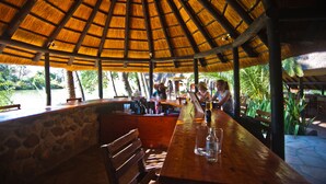 Un restaurante al aire libre con techo de paja, paredes de piedra y vistas a un lago.