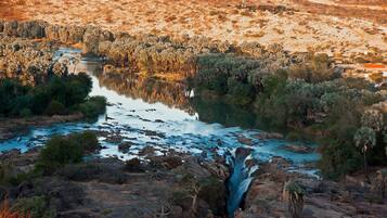 Een rivier met een waterval, omringd door schaarse begroeiing en enkele gebouwen op de achtergrond.