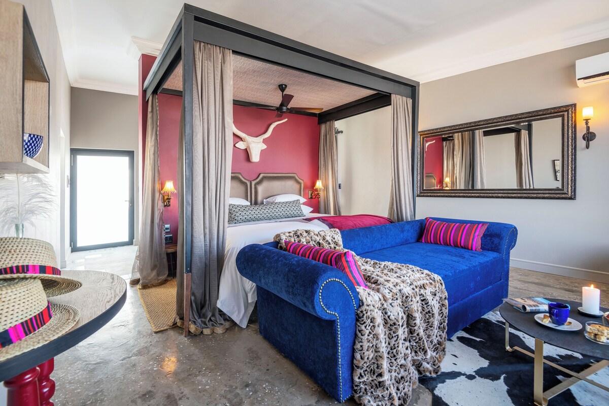 A bedroom with a four-poster bed, a blue sofa, a cow skull wall decoration, a mirror, and a small table with a candle.