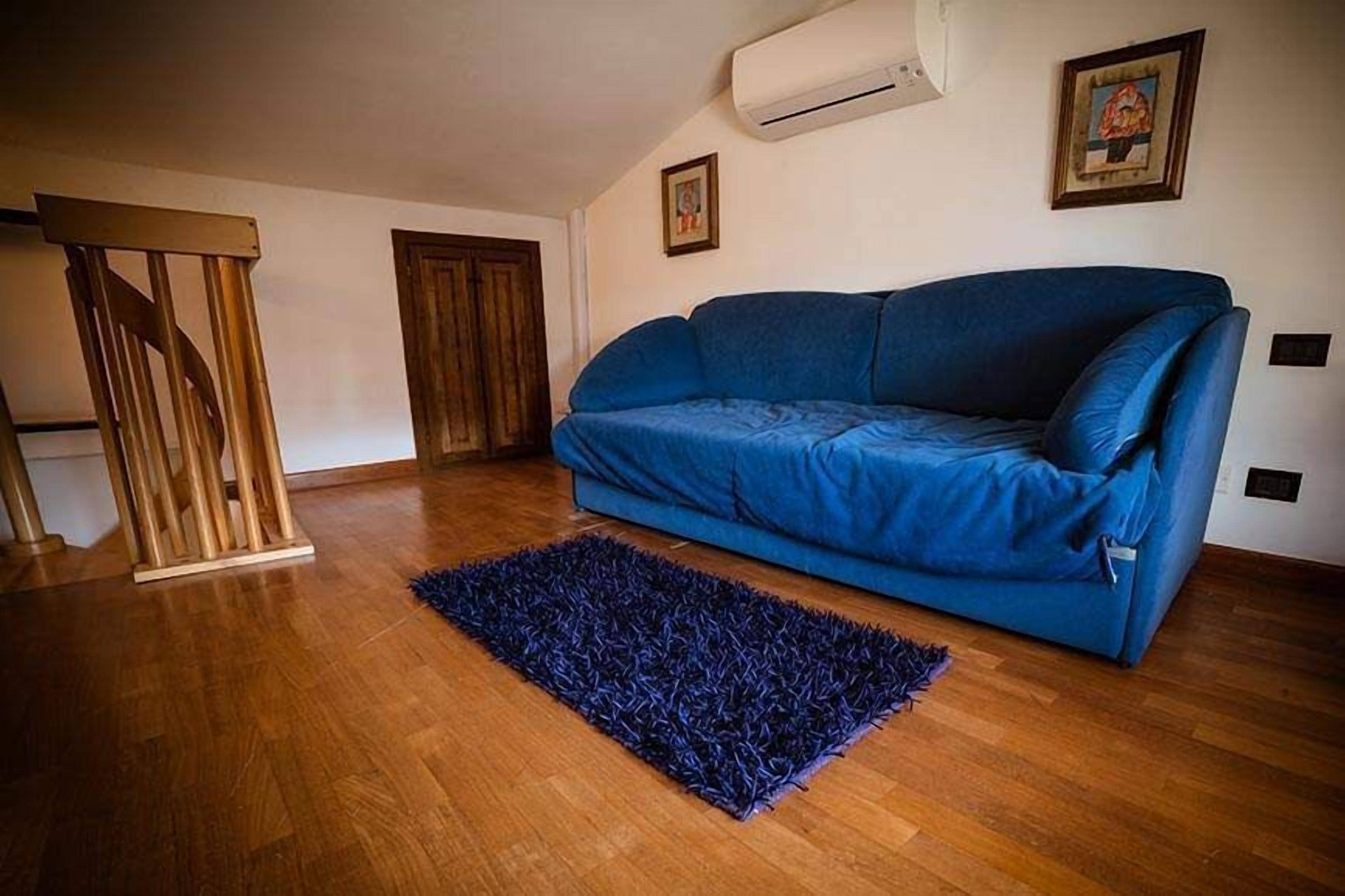 A room with a blue sofa, wooden flooring, a dark blue rug, a wooden chair, and an air conditioning unit.