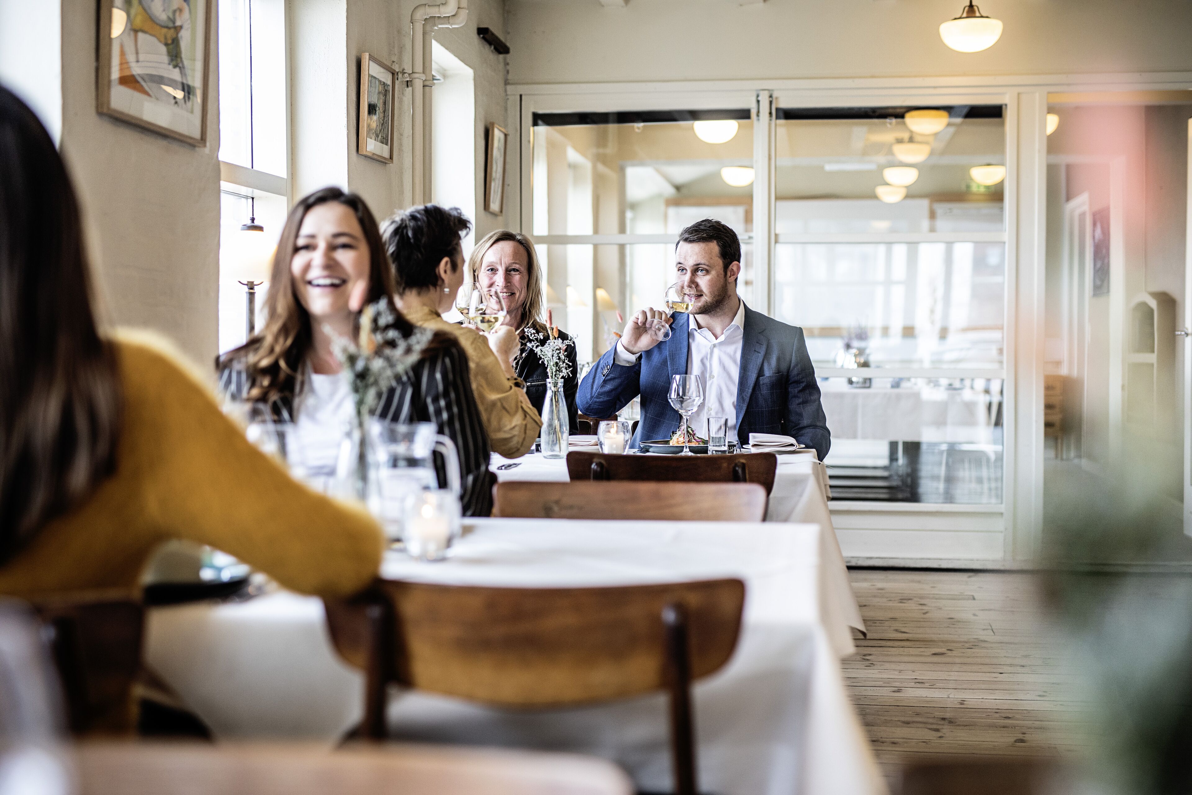 En restaurantindretning med træborde og stole, billeder i rammer på væggene og et stort vindue, der lukker naturligt lys ind.