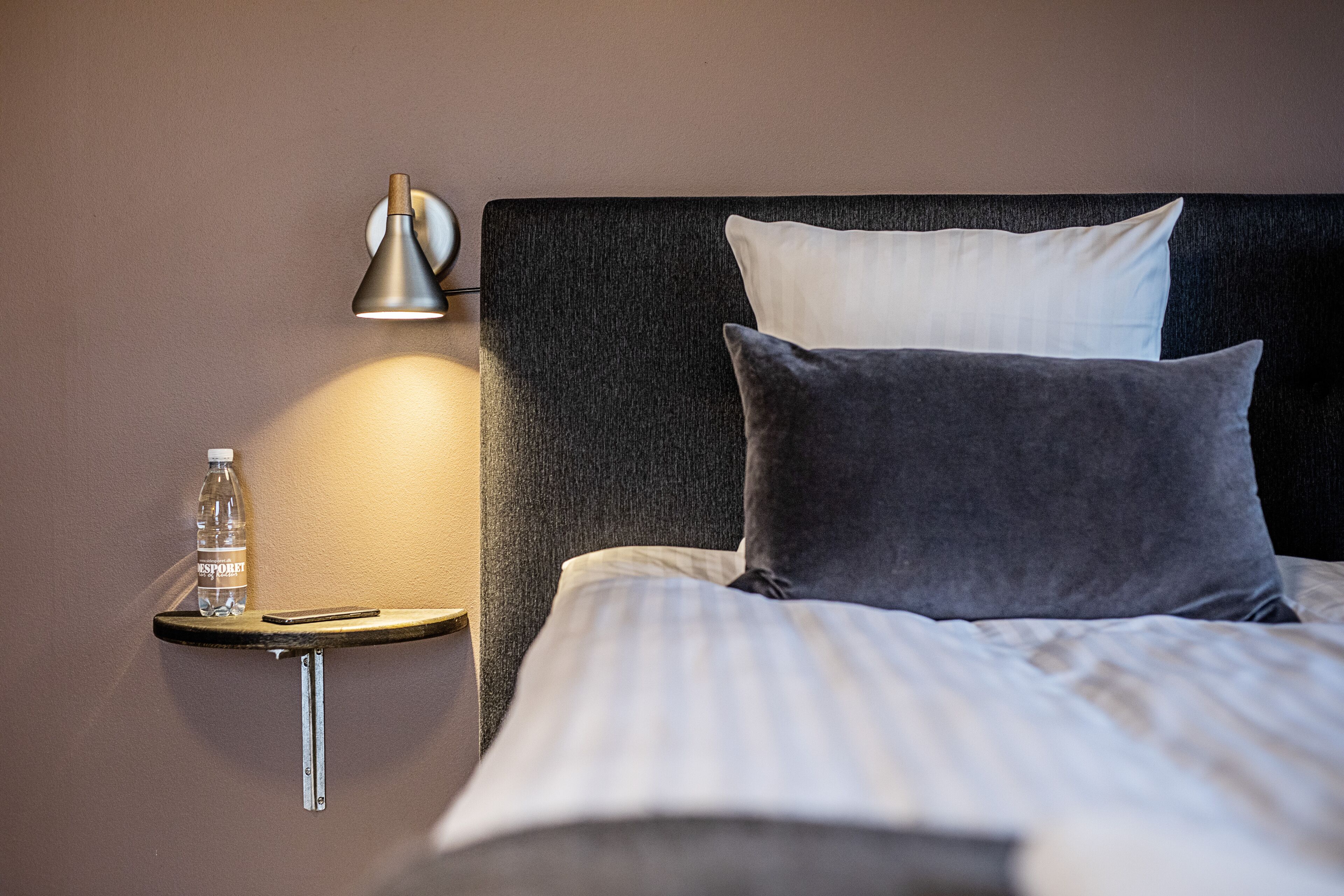 A neatly made bed with two pillows, a bedside table with a lamp, a bottle of water, and a smartphone.