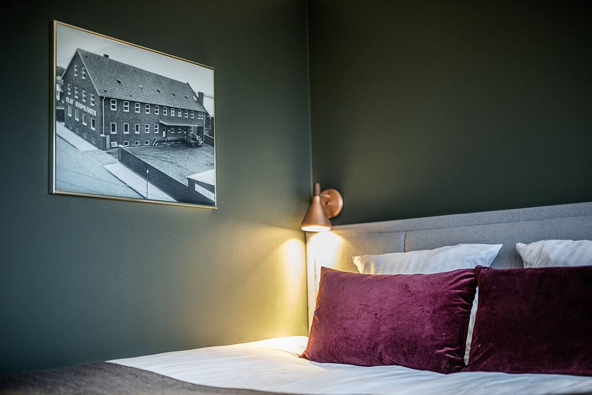 A neatly made bed with white linens and a single purple pillow, a framed black and white photo of a building on the wall, and a bedside lamp.