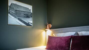 A neatly made bed with white linens and a single purple pillow, a framed black and white photo of a building on the wall, and a bedside lamp.