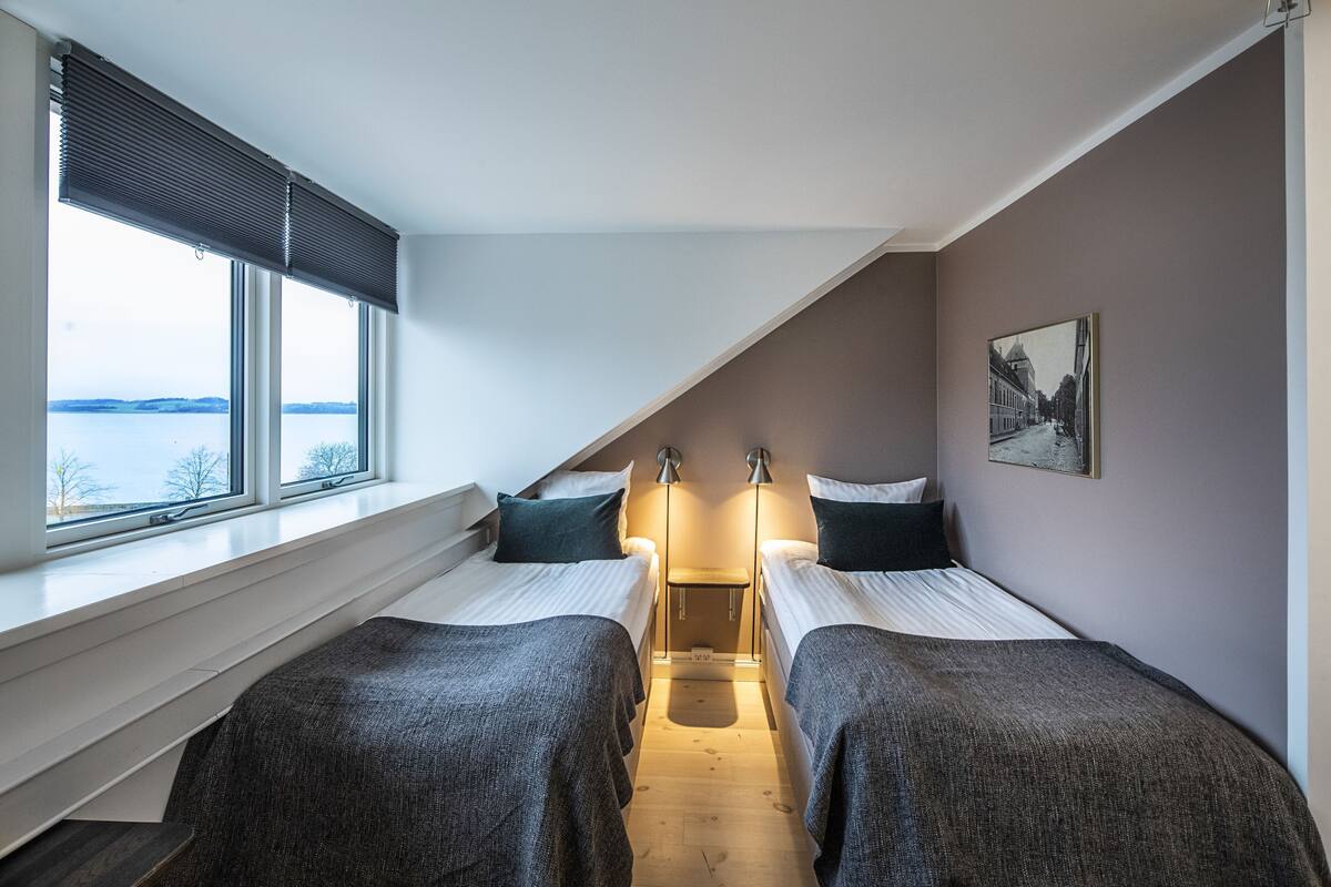 A bedroom with two beds, a window with blinds, a bedside table, and a wall-mounted picture.