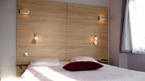Habitació d'hotel amb capçalera de fusta, dues làmpades de nit, un llit amb roba blanca i un coixí vermell, i una tauleta de nit amb un comandament a distància.