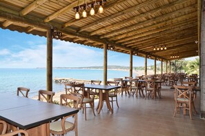 An outdoor dining area with wooden tables and chairs, overlooking a beach and the sea.