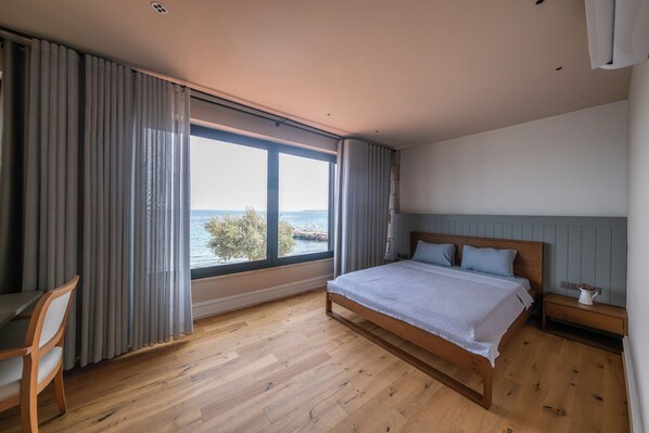 A bedroom with a large window, wooden floor, a bed with a headboard, a bedside table, and a chair.