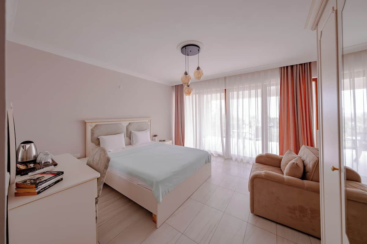 A modern hotel room with a large bed, a sofa, a desk with books and a kettle, and a view of the outdoors through sheer curtains.