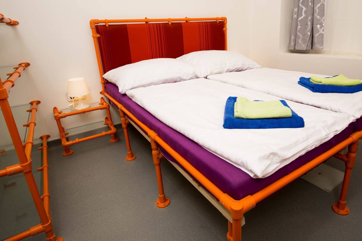 Ein Doppelbett mit Holzrahmen, einer violetten Matratze und weißer Bettwäsche. Neben dem Bett steht ein kleiner Tisch mit einer Lampe.