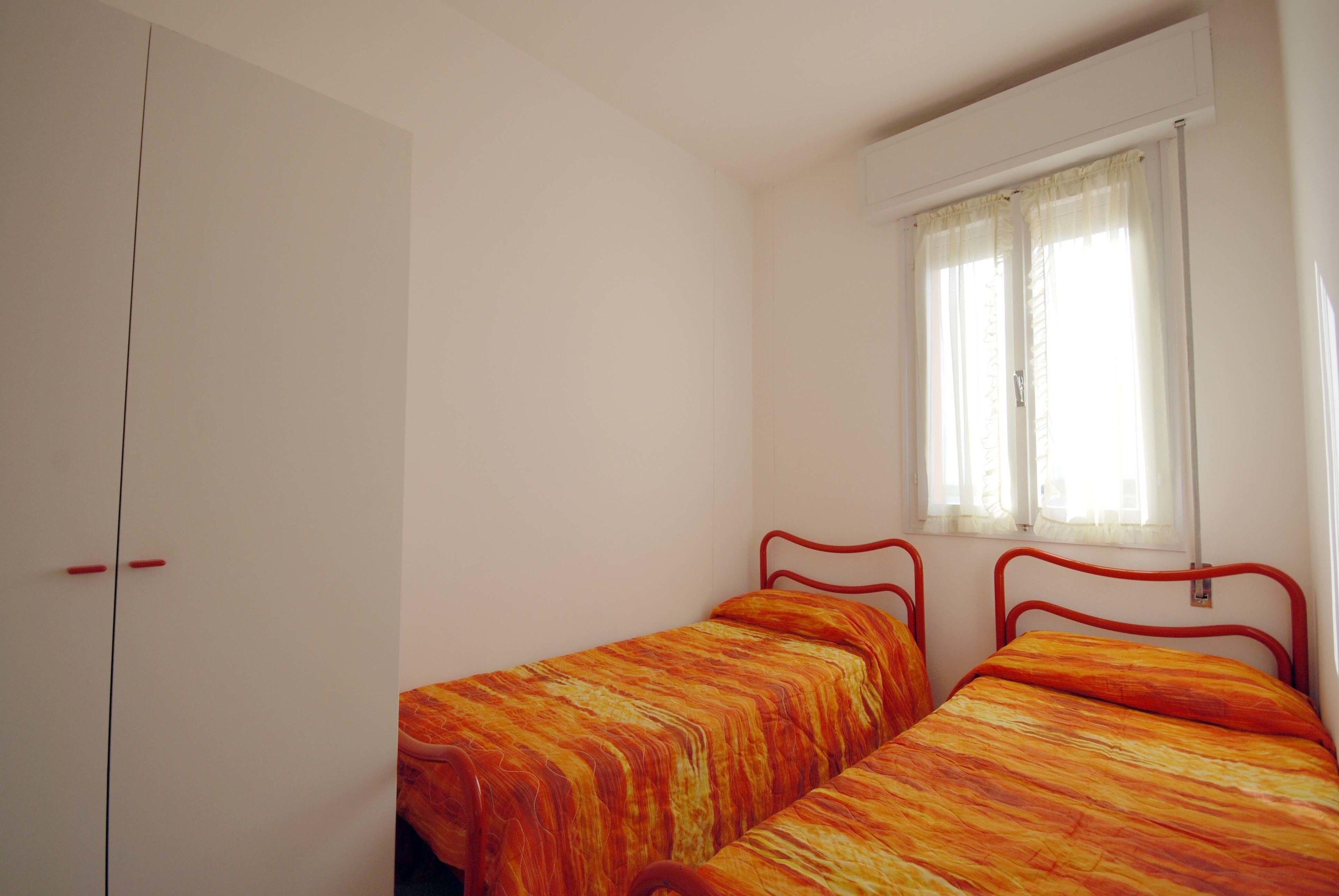 Quarto com duas camas de solteiro, um guarda-roupa e uma janela com cortinas.