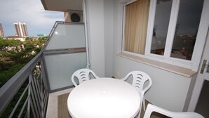 Een balkon met een ronde tafel en vier stoelen, een raam met gordijnen en uitzicht op gebouwen en bomen.