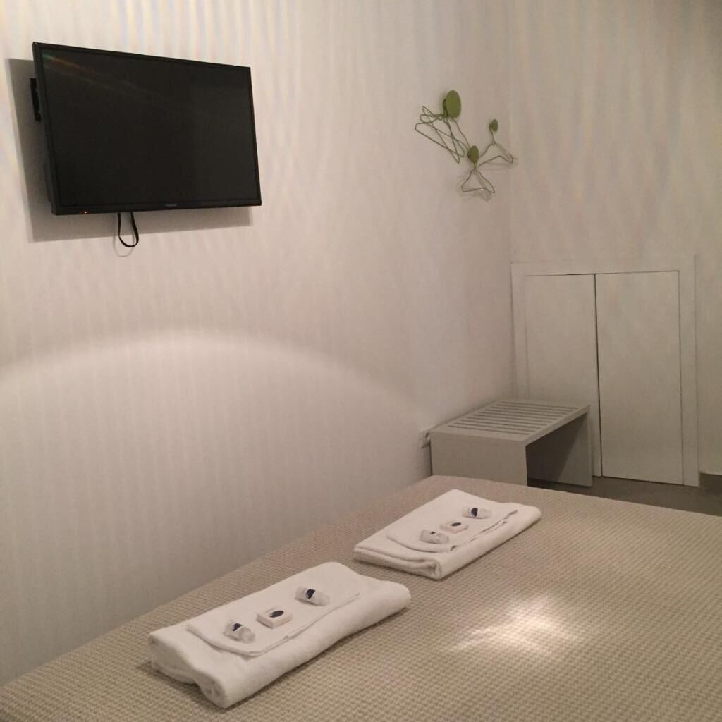 A hotel room with a flat-screen TV mounted on the wall, a small plant on the wall, a bench, and two neatly folded towels on the bed.
