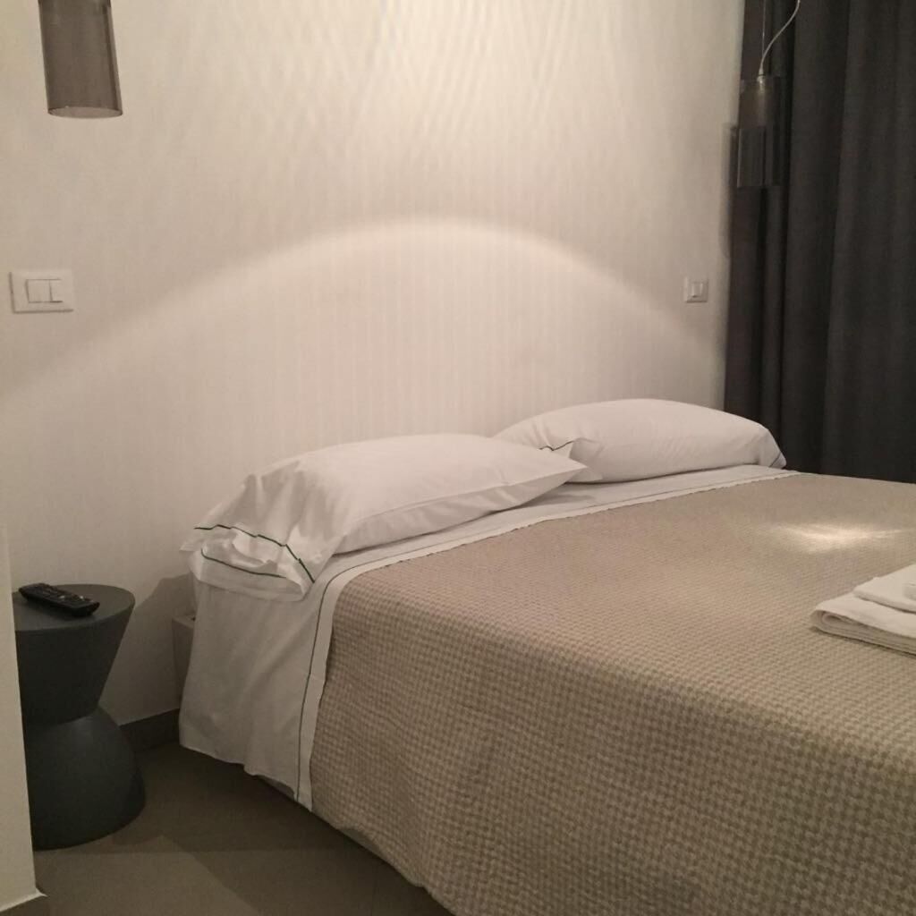 Un lit bien fait, avec du linge de lit blanc et une housse de couette beige à texture. Une télécommande est posée sur une petite table ronde, à côté du lit.