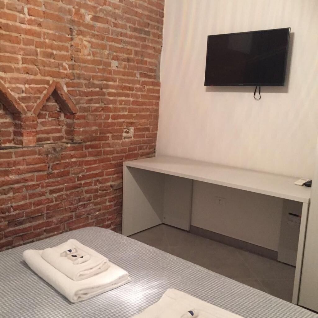 A room with a brick wall, a flat-screen TV mounted on the wall, a bed with a folded towel, and a bench.
