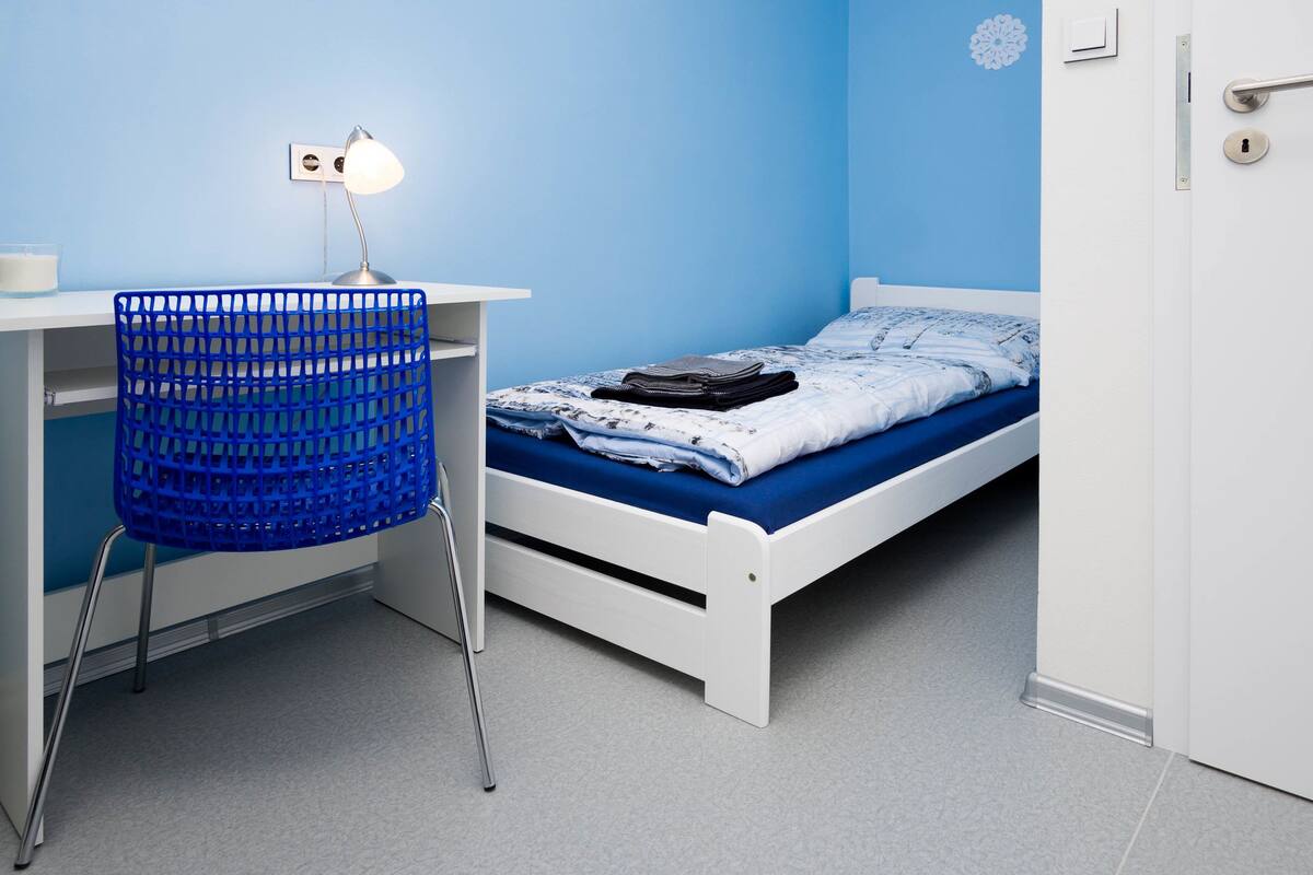 Een slaapkamer met een wit bed, een blauwe stoel, een bureau met een lamp en een deur met slot.