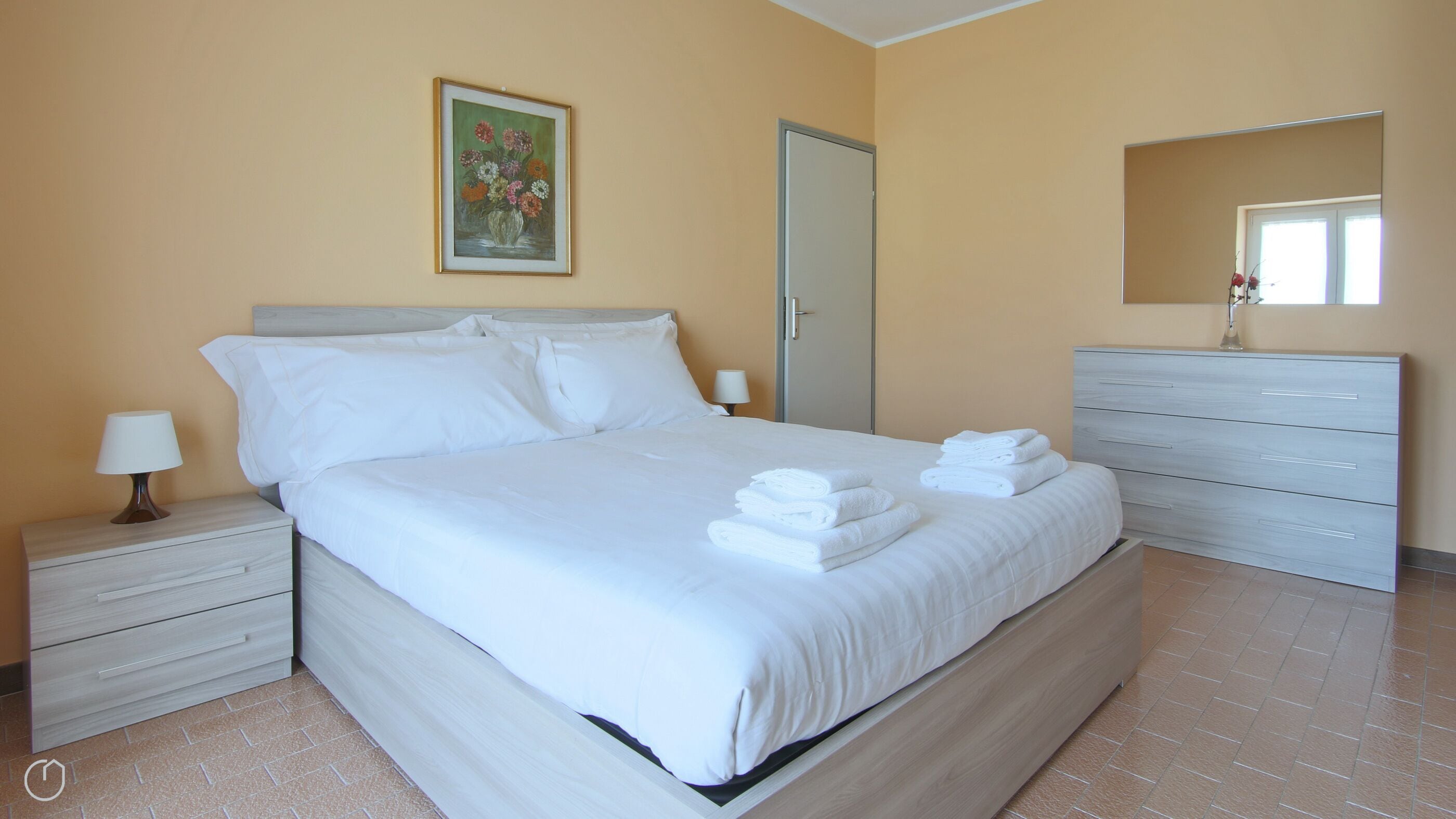 A neatly made bed with white linens and towels, a bedside table with a lamp, a dresser with drawers, and a painting on the wall.
