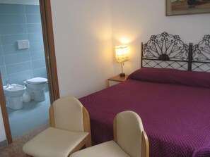 A bedroom with a bed, two chairs, a bedside table with a lamp, a bathroom with a toilet and sink, and a wall-mounted light fixture. - B&B Samuel (Verona)