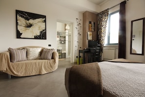 A bedroom with a bed, a sofa, and a desk. There is a window with curtains, a painting on the wall, and a radiator. - Ca' Bianca Bed & Breakfast (Fosdinovo)