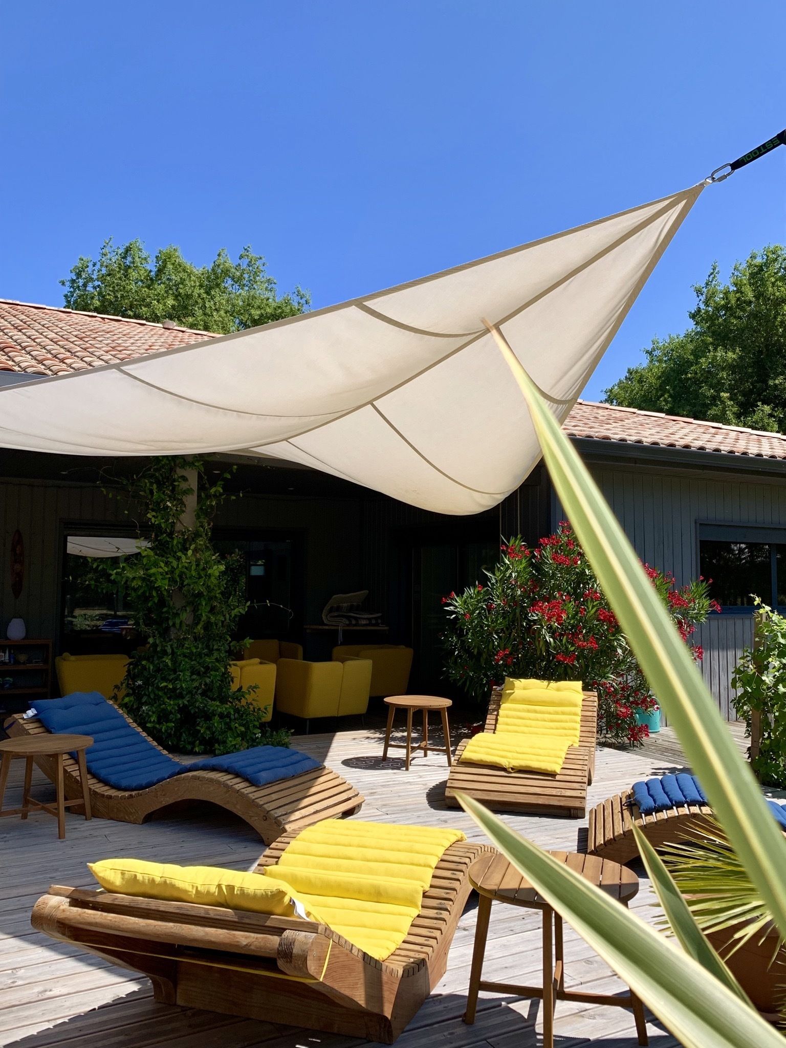A wooden deck with lounge chairs and a table, a large shade sail, and a building with a tiled roof in the background.