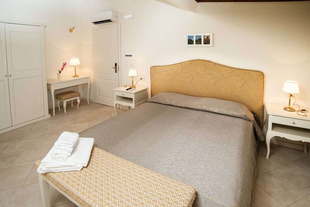 A neatly arranged bedroom with a large bed, a bedside table with a lamp, a small desk with a lamp, a wardrobe, and a bench with towels.