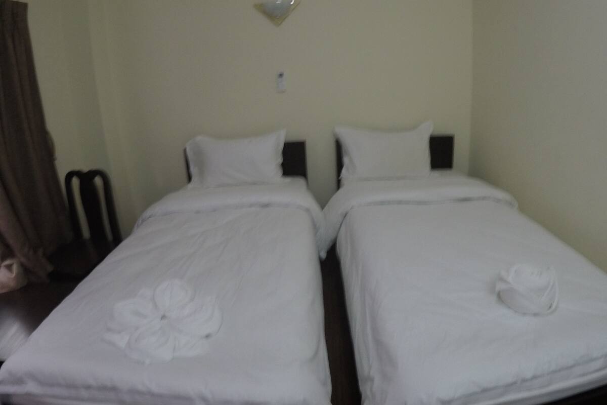Habitación de hotel con dos camas individuales, cada una con una colcha blanca y una almohada con motivos florales. Hay una mesita con una silla, y una lámpara de pared sobre las camas.