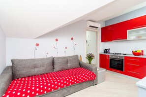 A modern living room with a grey sofa and a red polka dot throw blanket. Adjacent to the living room is a kitchen with red cabinets and a black oven.