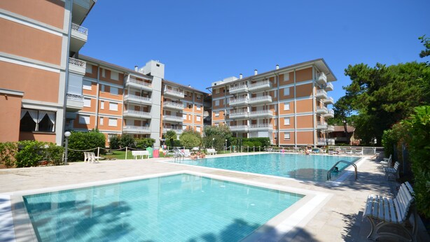 A swimming pool area with a clear blue pool, surrounded by a multi-story building with balconies, and a few lounge chairs. - Residence Gardenia (Lignano Sabbiadoro)
