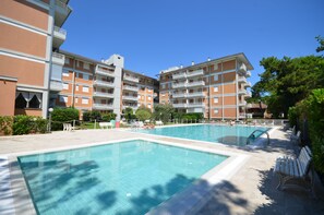Un espace piscine avec une piscine à l’eau d’un bleu limpide, entouré d’un immeuble à plusieurs étages doté de balcons, et agrémenté de quelques chaises longues.