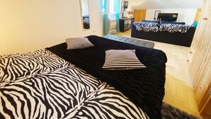 Une chambre à coucher avec une couvre-lit à motifs zébrés, une grande fenêtre avec des rideaux, une télévision et un petit meuble.