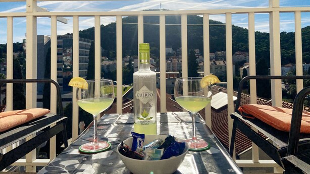 Un balcon avec une table dressée pour deux personnes, agrémentée d’une bouteille de spiritueux, de deux verres et d’un bol de collations.