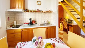 Une cuisine avec des armoires en bois, un évier, une cuisinière et une table à manger avec des chaises. Il y a une horloge au mur et un ordinateur portable sur un bureau dans la pièce adjacente.