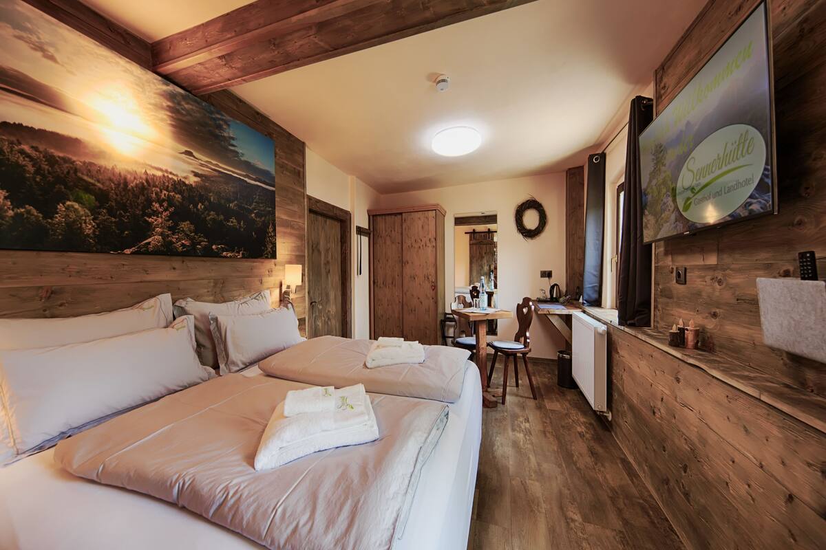 A bedroom with a large bed, wooden walls, a painting of a sunset, a desk with a computer, and a sign advertising a summer event.
