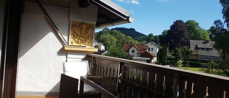 A balcony with wicker furniture, a white wall with a decorative panel, and a view of houses and trees in the background.