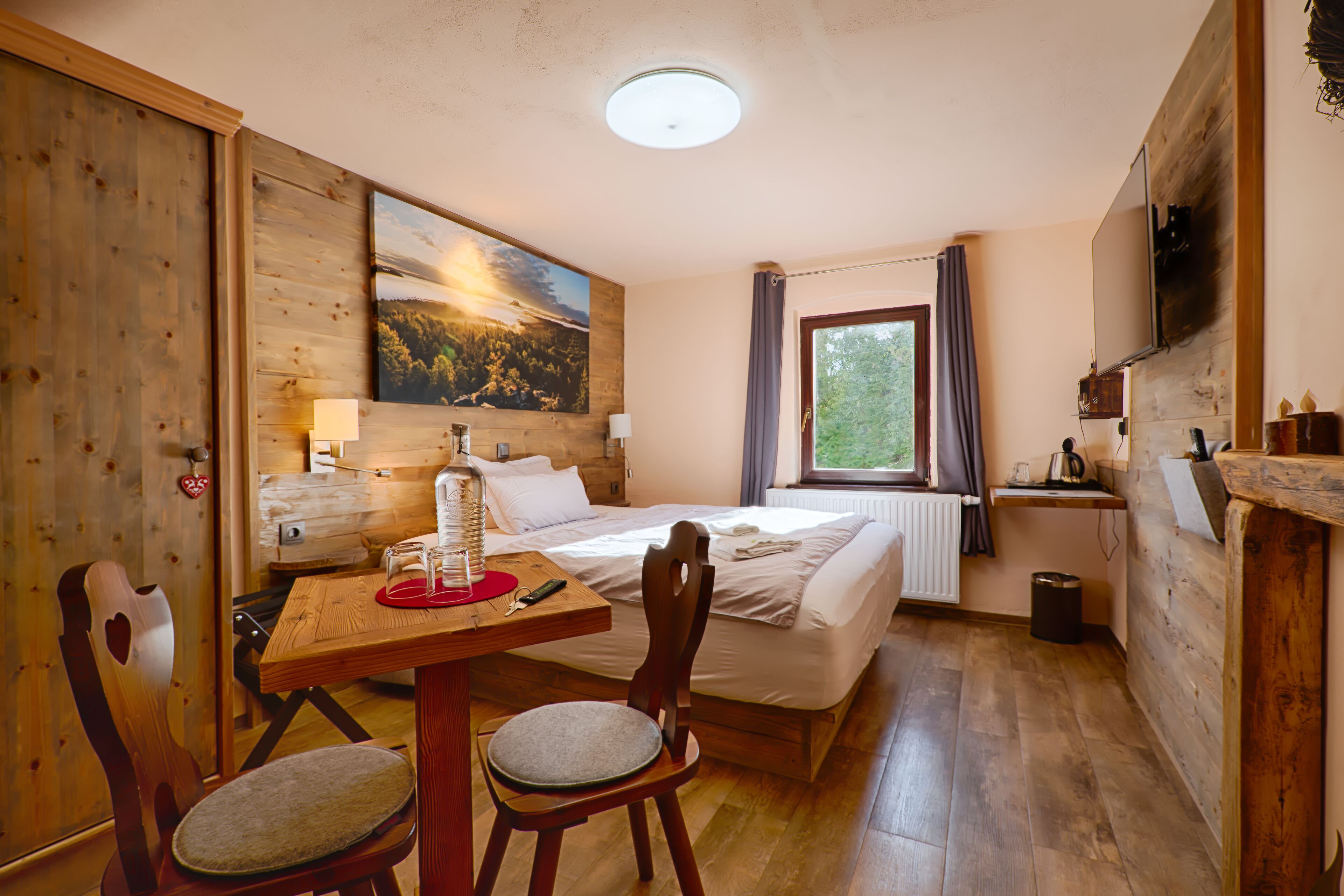 A cozy room with a wooden floor, a bed with white linens, a wooden table with chairs, a painting on the wall, and a window with curtains.