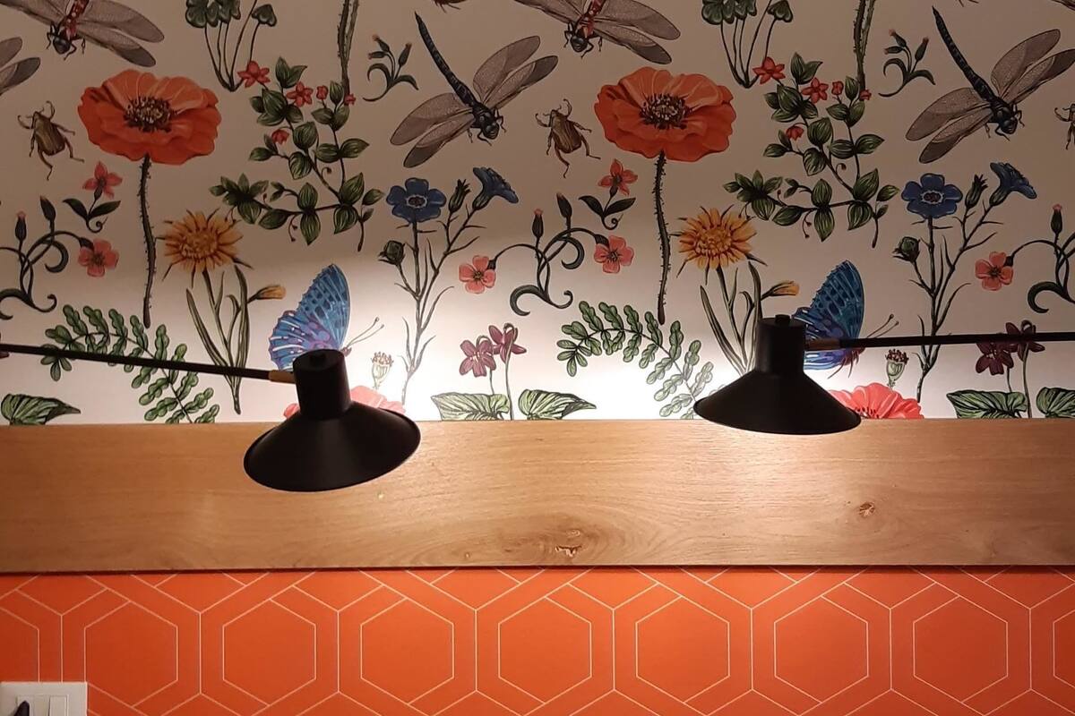 A room with a floral wallpaper, a wooden shelf with two black lamps, and a green bed with a white towel.