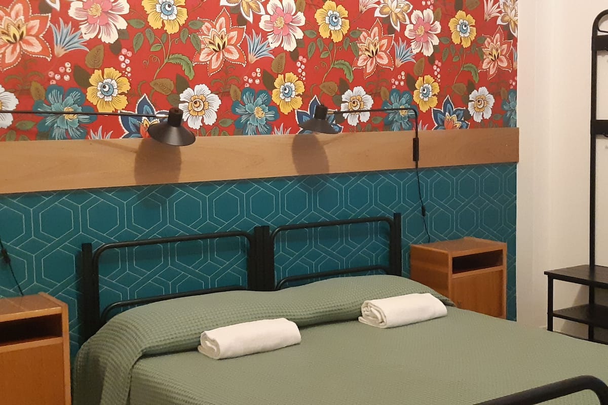 A neatly made bed with a green bedspread and two rolled towels, a wooden nightstand with a lamp, and a wall with a floral pattern.