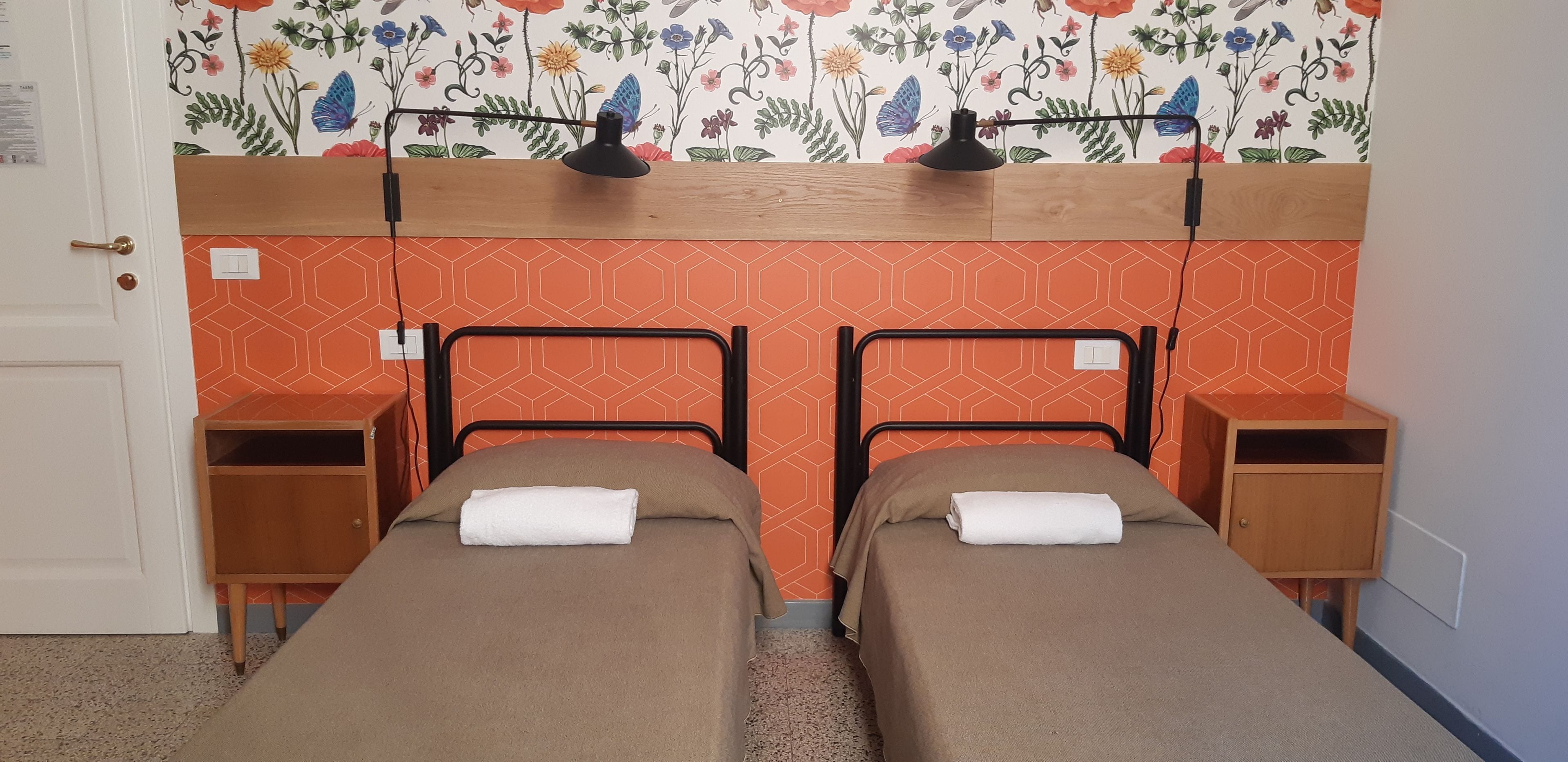 A room with two beds, each with a wooden nightstand, a lamp above, and a floral wallpaper.