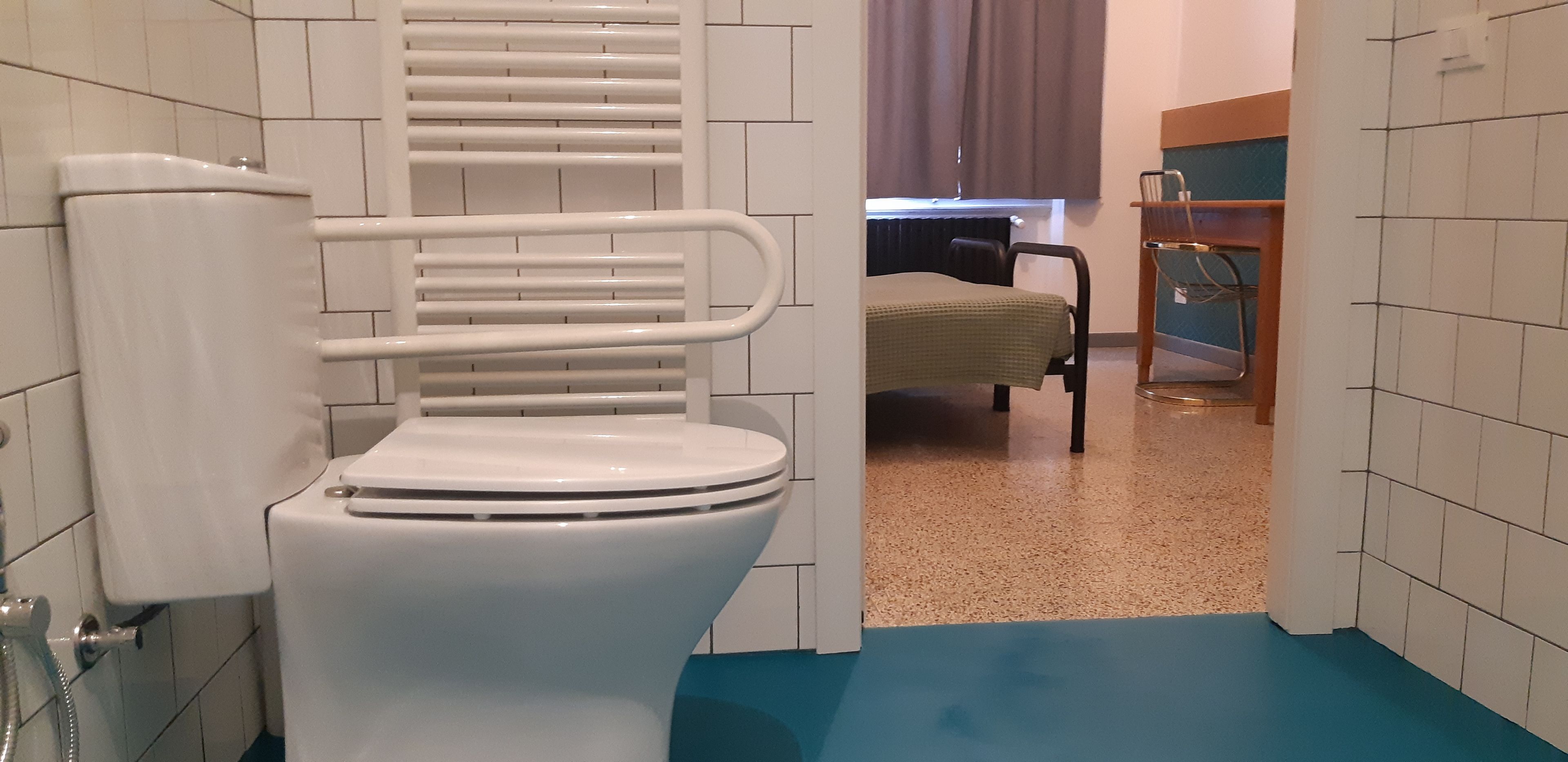 A bathroom with a white toilet, a shower area, and a bed in the adjacent room.