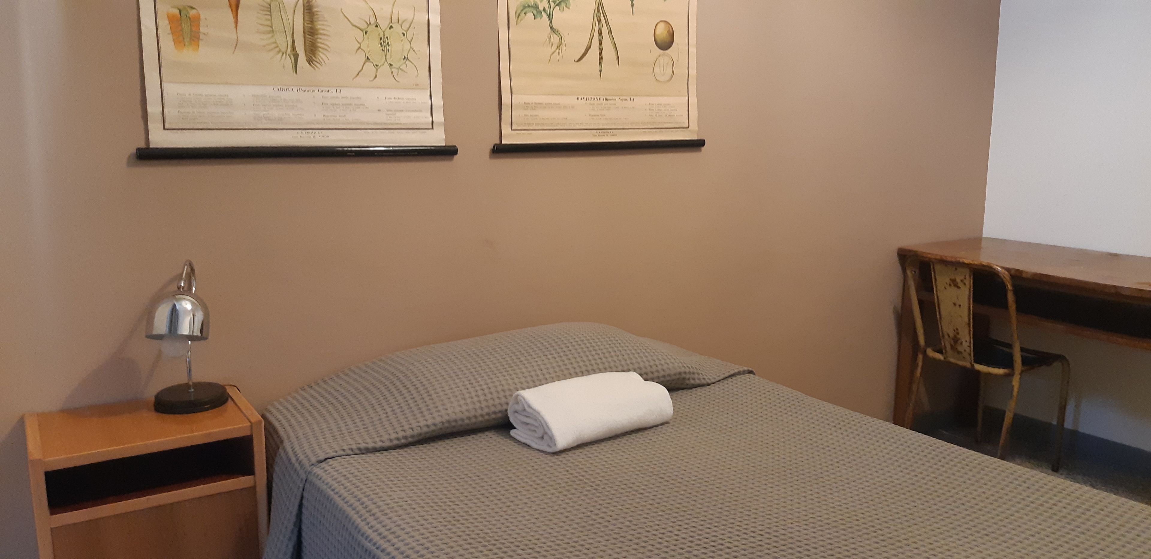 A neatly made bed with a folded towel, a small wooden nightstand with a lamp, and two framed botanical illustrations on the wall.