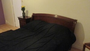 Een slaapkamer met een houten bed, een donkerdekkens, een nachtkastje met een bloevas en een houten stoel.