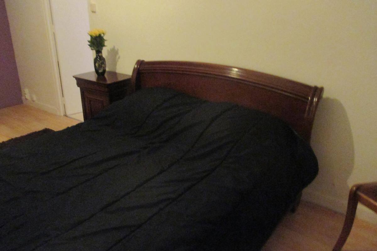 Een slaapkamer met een houten bed, een donkerdekkens, een nachtkastje met een bloevas en een houten stoel.